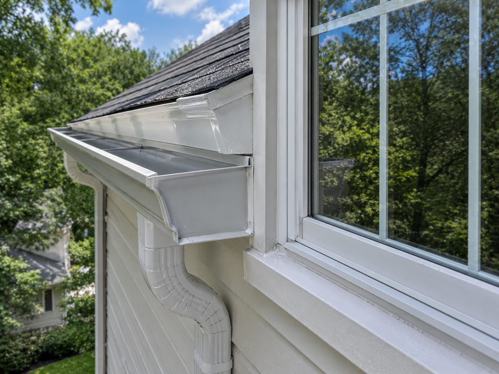 Close view of a freshly cleaned aluminum gutter and downspout on a suburban home with streak free exterior window glass visible beside it reflecting green foliage