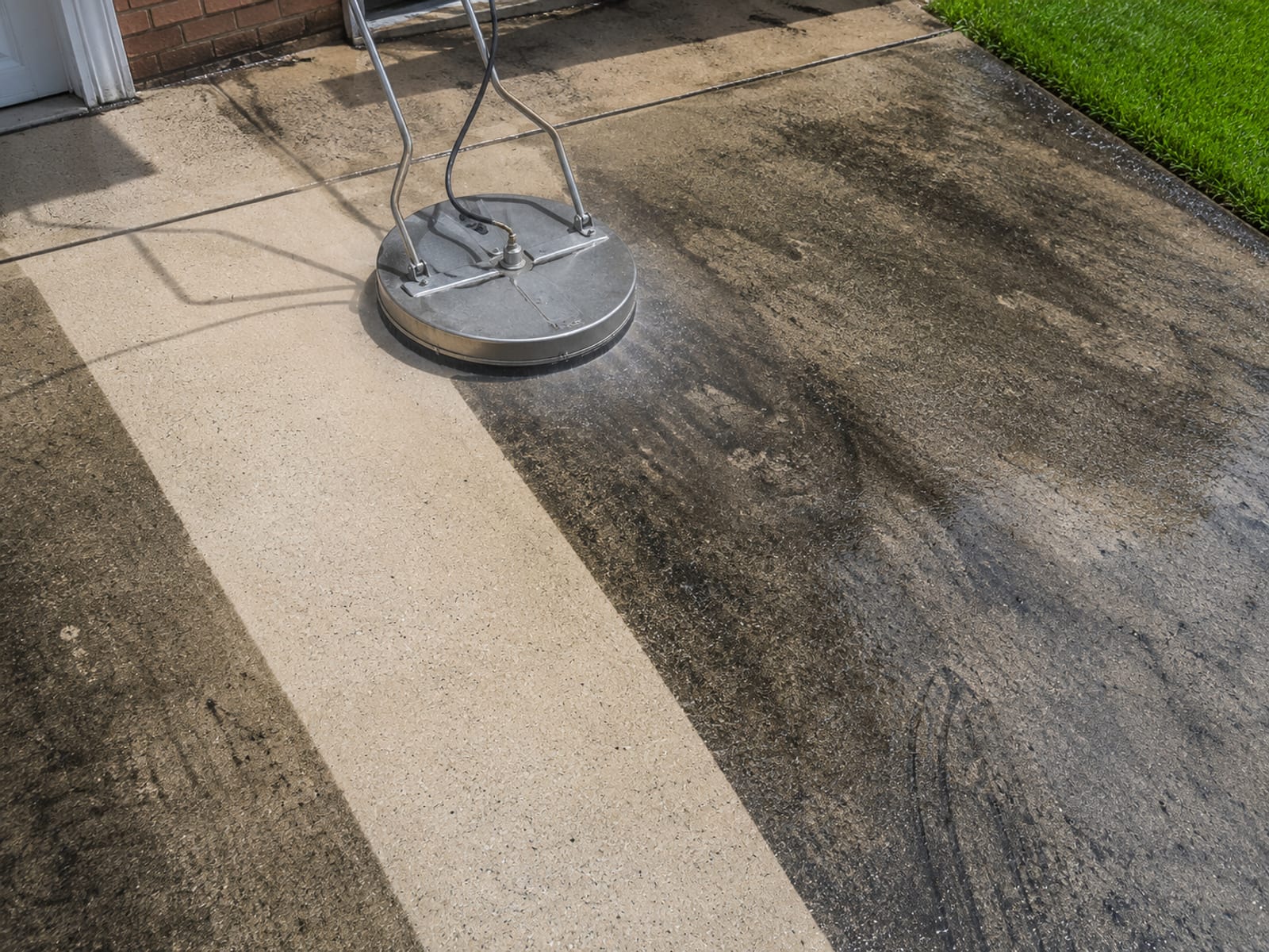Overhead angle of a concrete driveway being cleaned with a round surface cleaner attachment showing a stark clean path cut through black algae and tire stained concrete