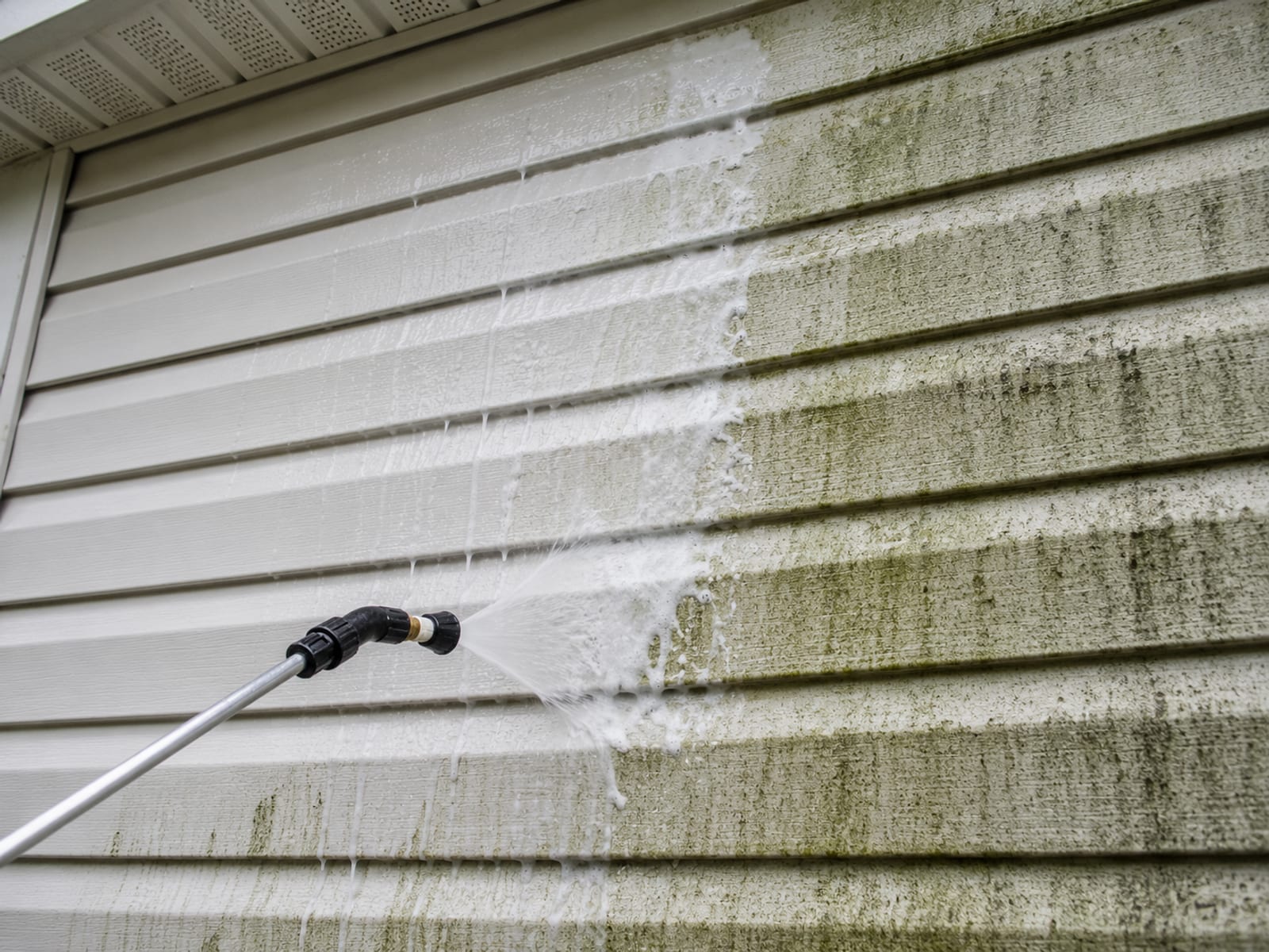 Close detail of a soft wash wand spraying white cleaning solution onto green algae streaked vinyl siding on a suburban home