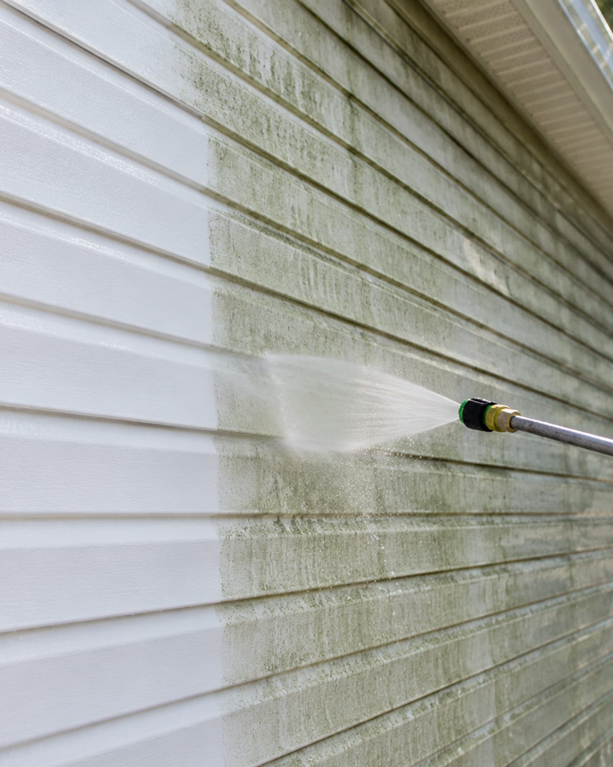 Angled close detail of a residential vinyl siding panel on a Baltimore County home showing one cleaned bright white section beside an algae streaked green grey dirty section with a pressure wash wand extended into frame