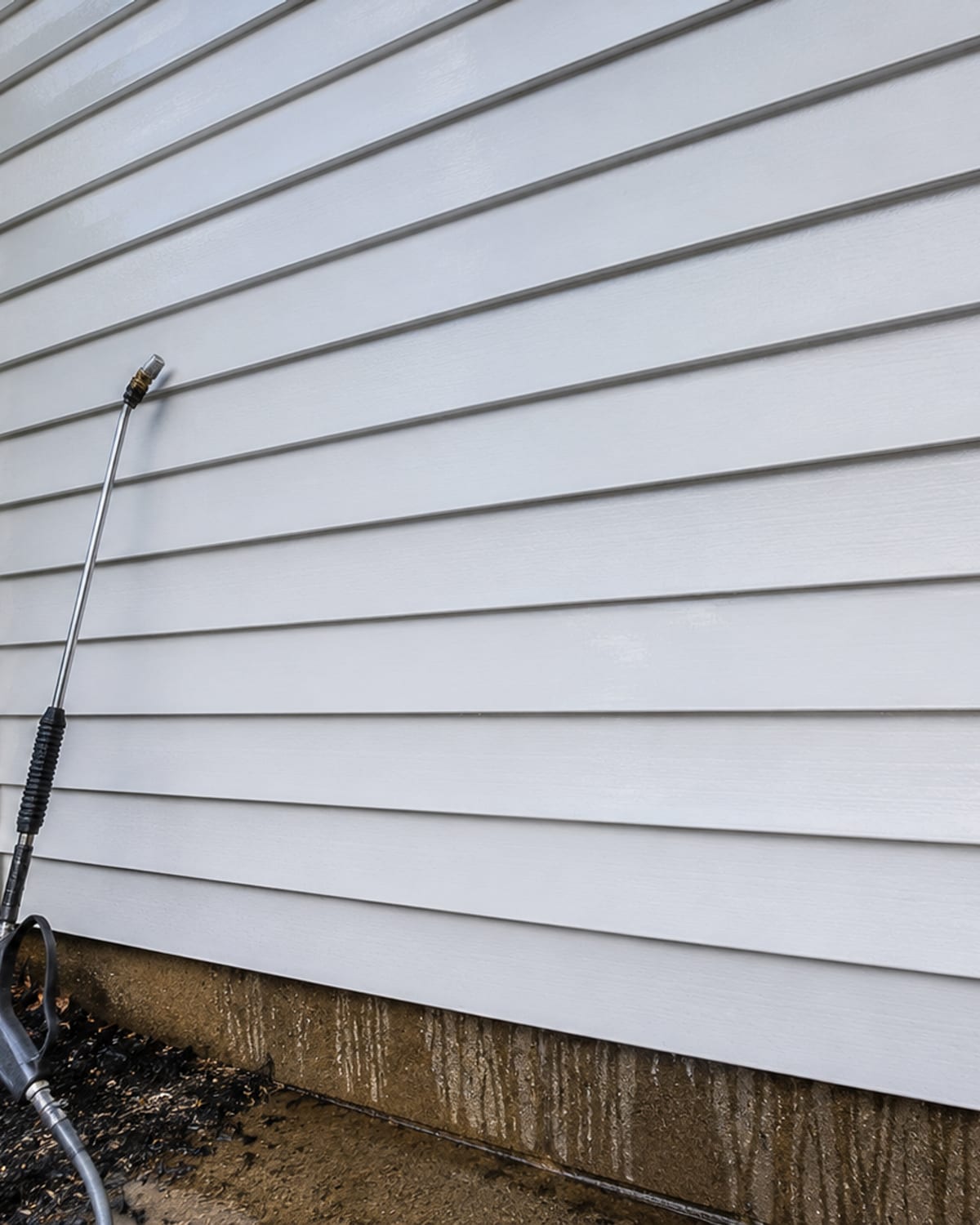 Close detail of a white vinyl siding wall on a Baltimore County colonial home showing streak-free clean panels with a pressure washing wand resting nearby and wet runoff visible at the base