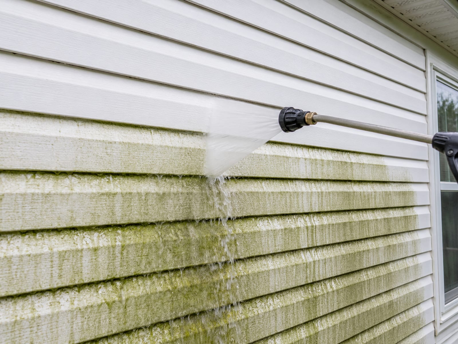 Close angle view of a pressure washing wand and wide fan nozzle directed at green algae stained white vinyl siding on a suburban Perry Hall Maryland home