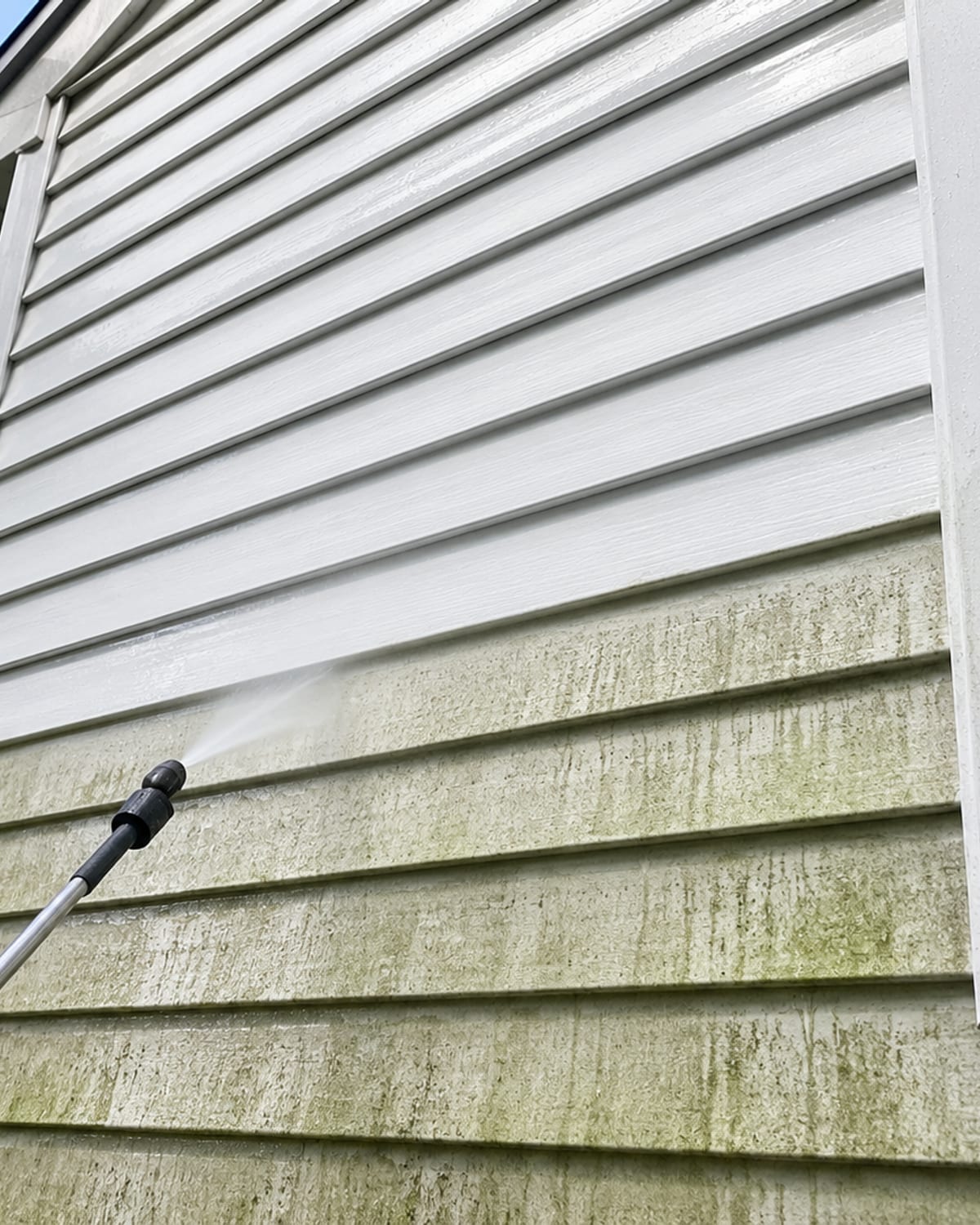 Close detail of white vinyl siding on a Maryland colonial home being cleaned with a soft wash wand showing clean bright panels above and algae stained green panels below